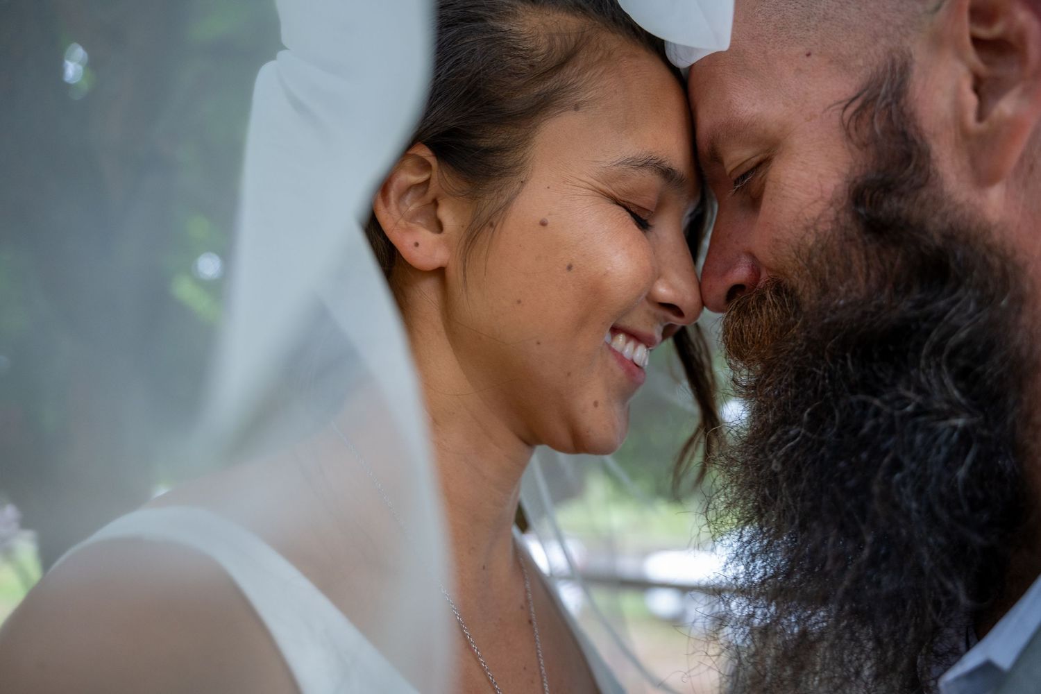 A smiling bride and bearded groom share an intimate forehead touch under a wedding veil outdoors.