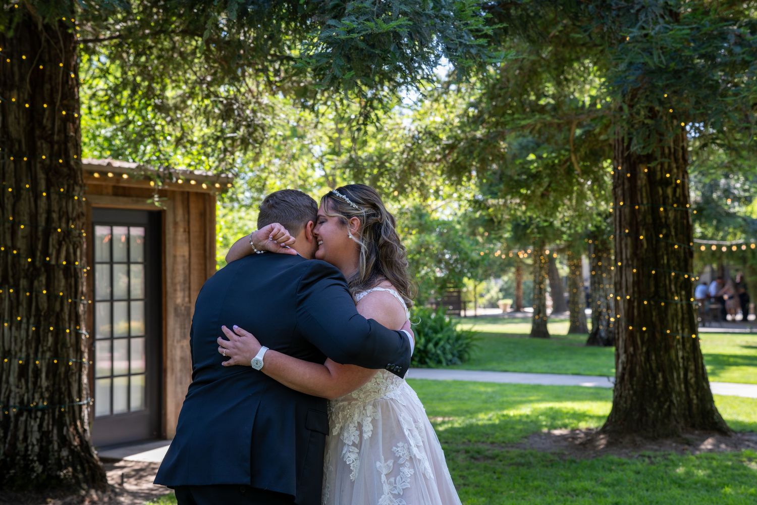 Bride and groom share first kiss surrounded by tall trees with fairy lights at outdoor wedding venue.