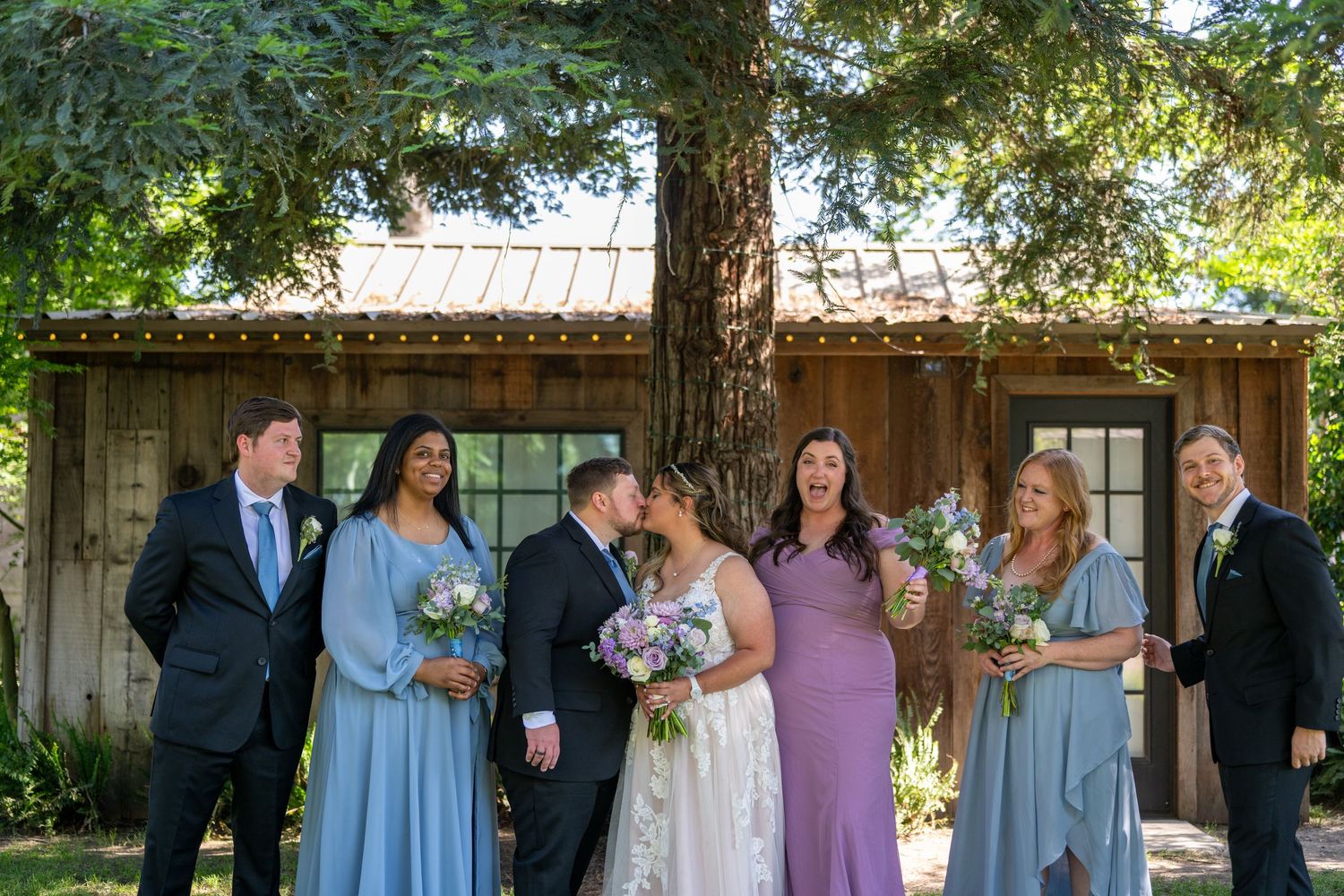 Wedding party poses outdoors near rustic barn, bride and groom kissing while bridesmaids in blue and purple hold bouquets.