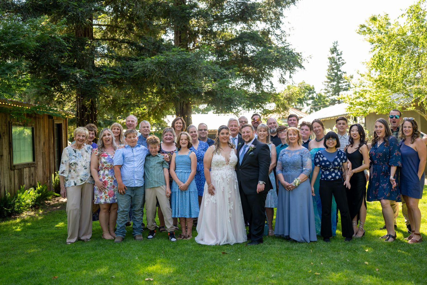 Large wedding party group photo outdoors on green lawn with trees, bride in white gown surrounded by guests in blue attire.
