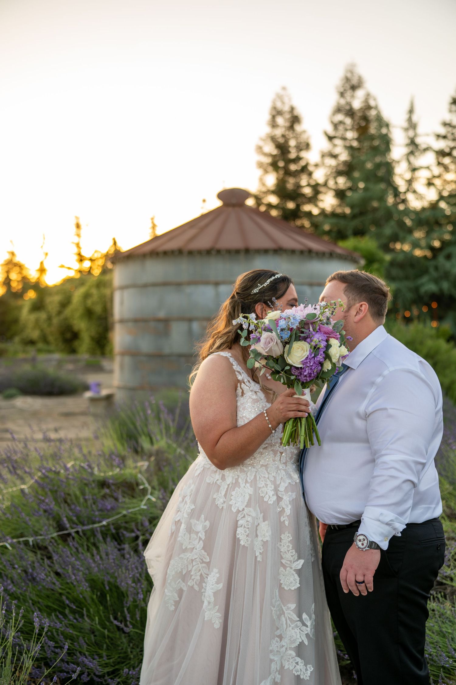 Bride and groom share a romantic moment at sunset, surrounded by lavender fields and tall trees at an outdoor wedding venue.