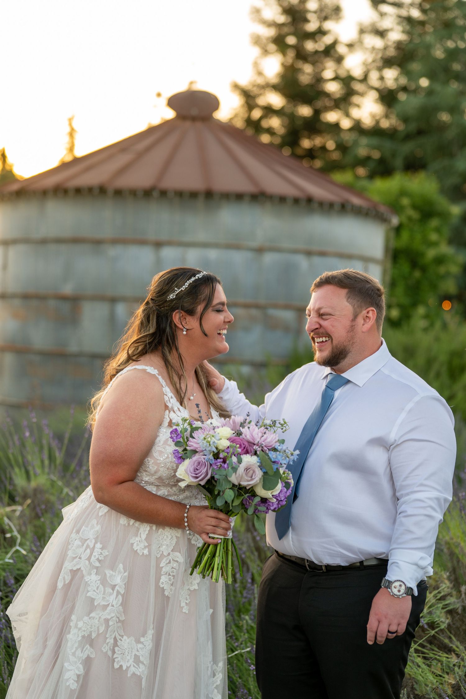 Bride and groom laughing together outdoors at golden hour, with a rustic grain silo and lavender garden backdrop.