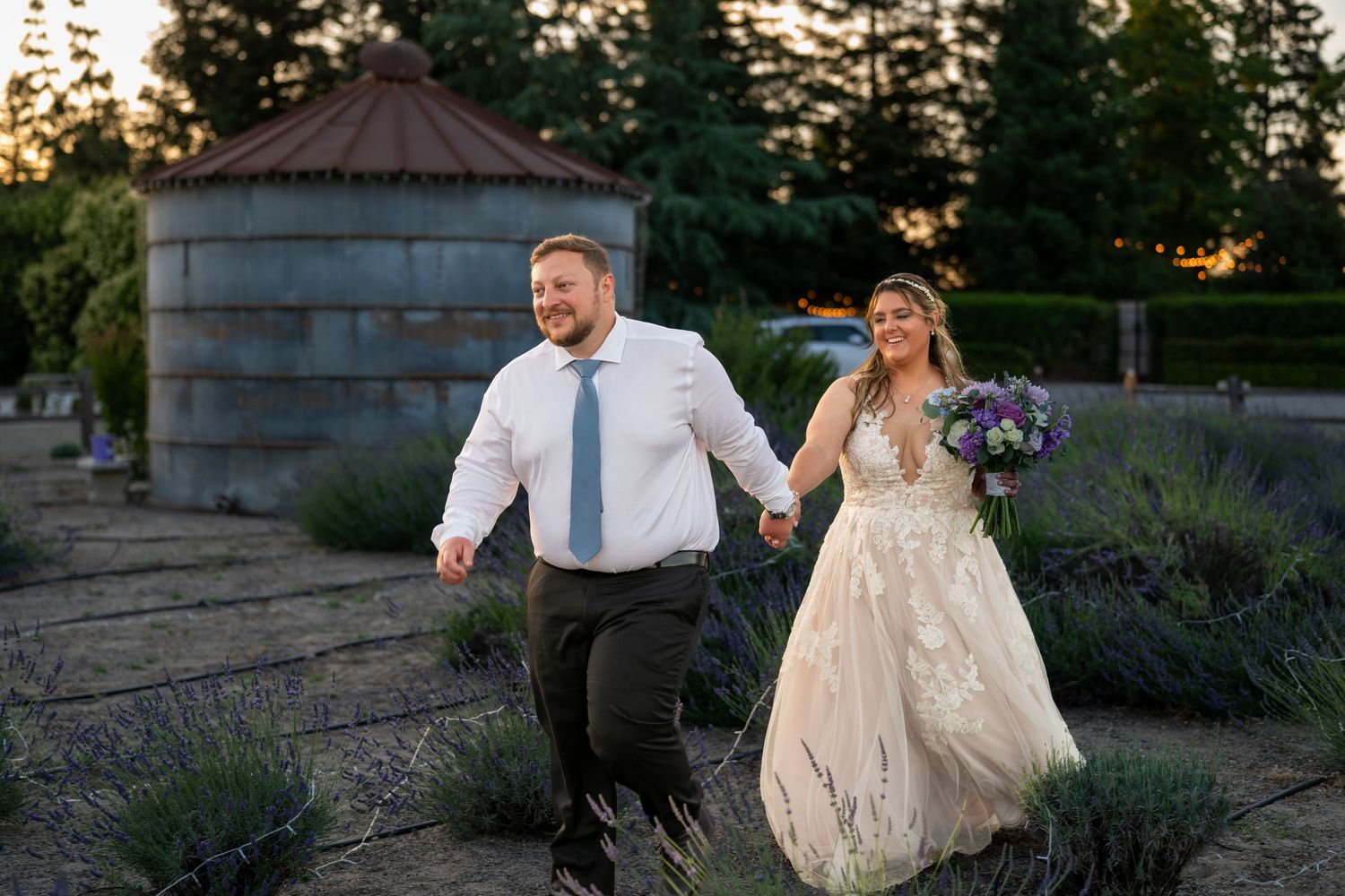 Bride and groom walk hand-in-hand through lavender fields at sunset, bride in lace gown holding purple bouquet.