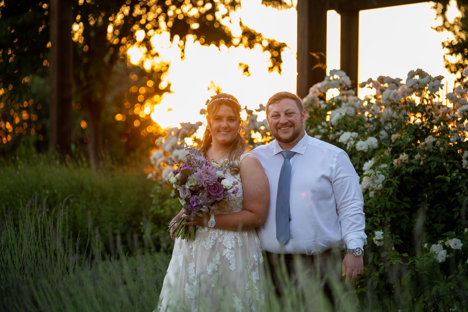 Bride and groom pose together at golden hour sunset surrounded by lush greenery and white flowers at outdoor wedding.