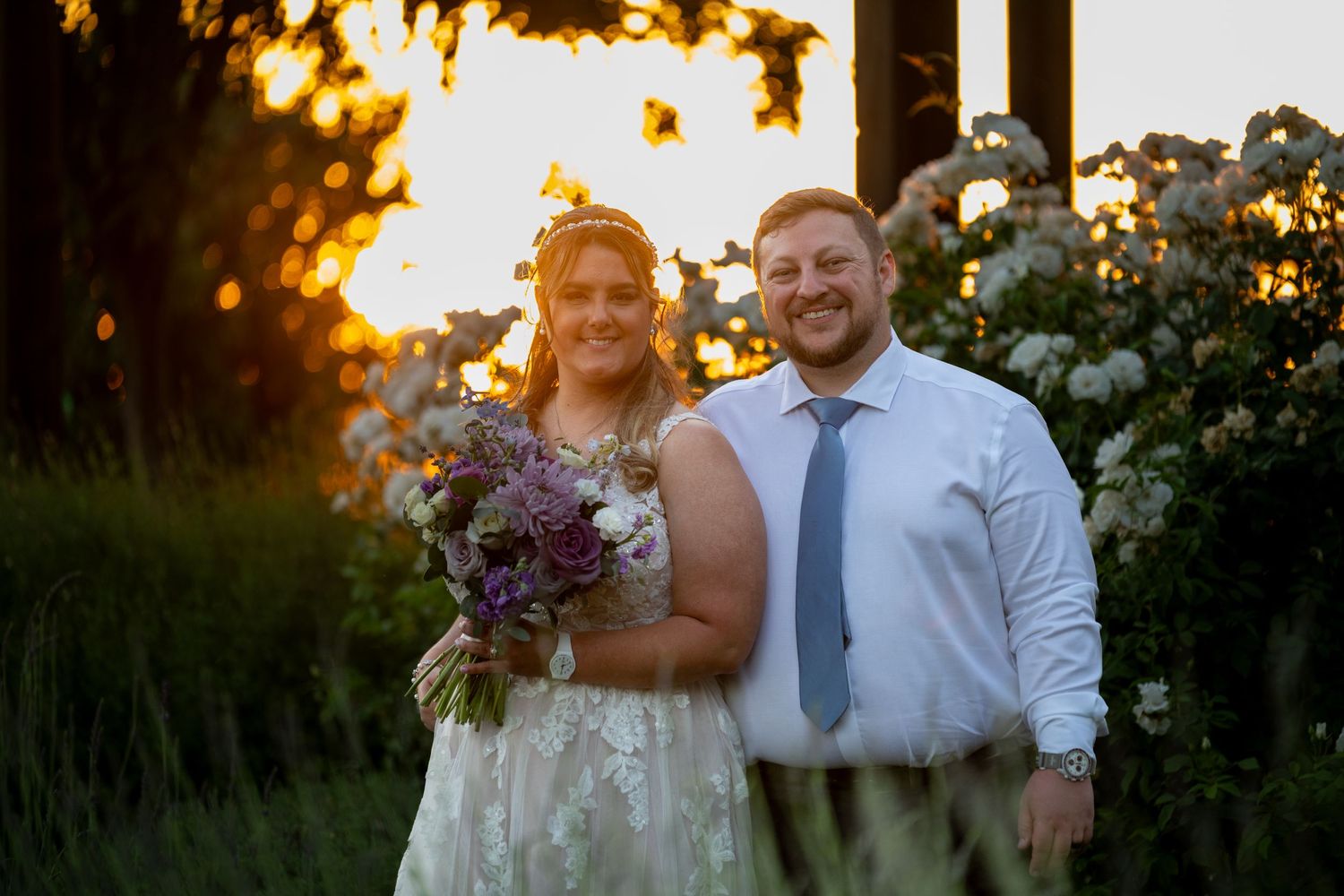 Bride and groom smiling at sunset surrounded by lush greenery and white flowers, bride holding purple bouquet.