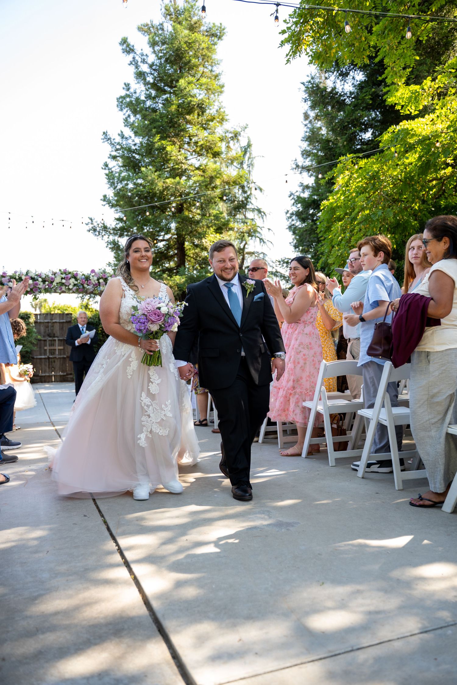 Bride in white gown holding purple bouquet walks down outdoor aisle with escort, guests watching at sunny wedding ceremony.