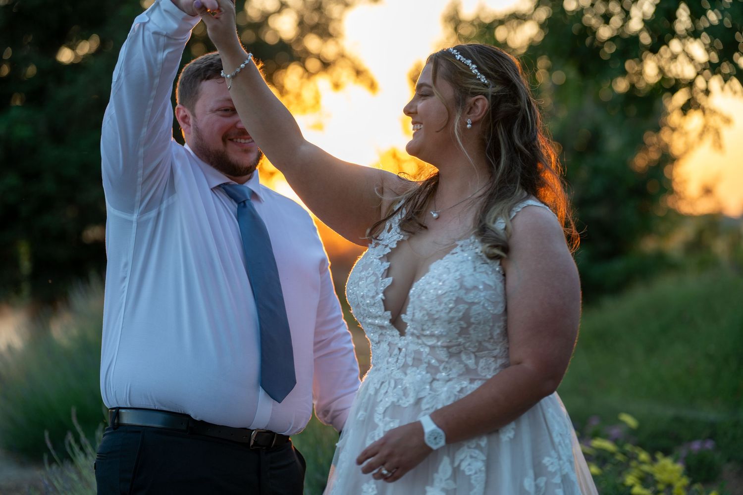 Bride in lace gown and groom in white shirt dance outdoors at golden hour sunset wedding celebration.