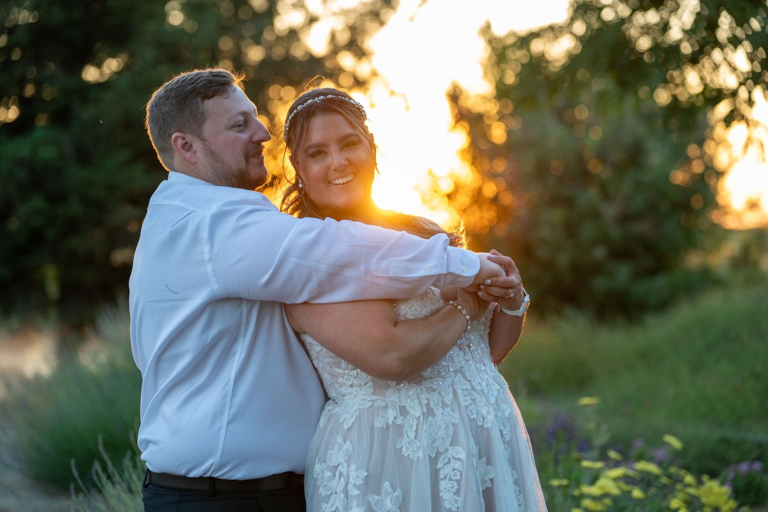 Couple dancing at golden hour sunset, bride in lace gown, groom in white shirt, surrounded by lush greenery.
