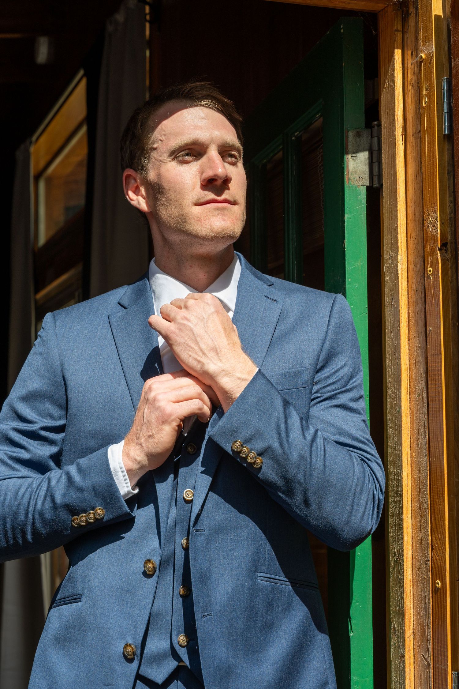 Man in blue three-piece suit adjusting tie outdoors in sunlight.