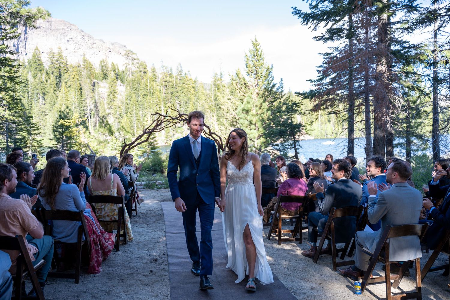 Newlywed couple walks down outdoor wedding aisle surrounded by guests, pine trees, and a mountain lake backdrop.