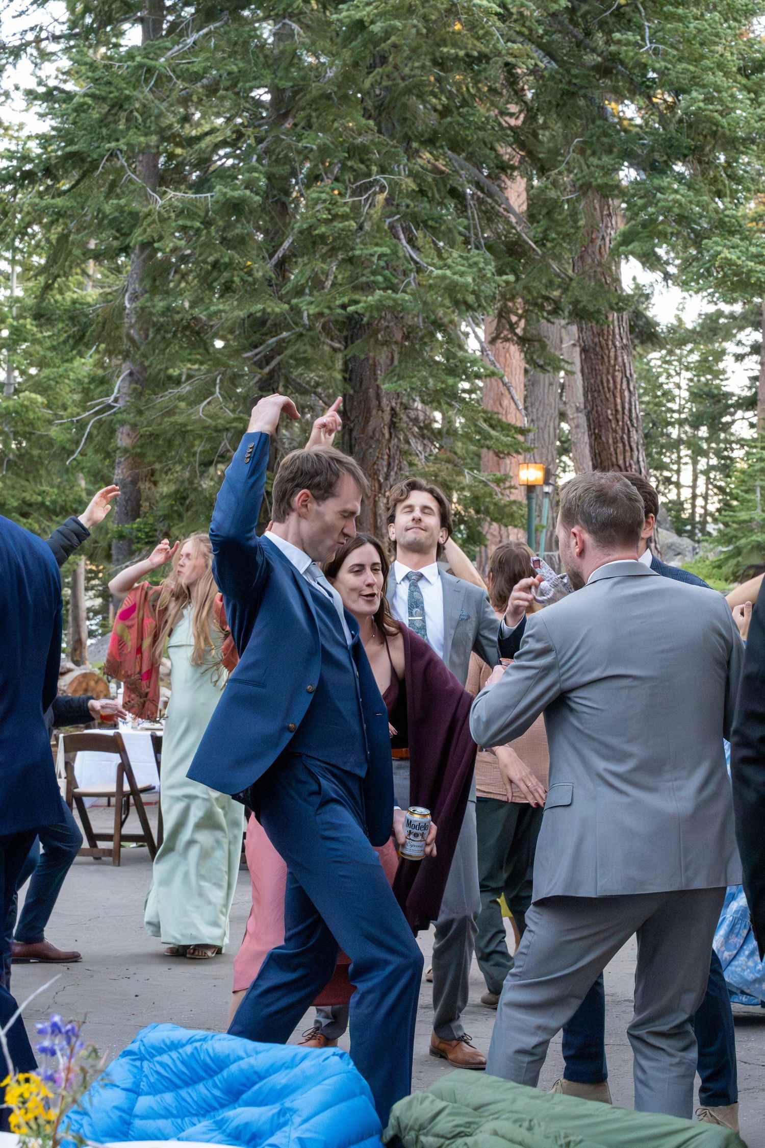 Wedding guests dancing outdoors among tall pine trees at evening reception.