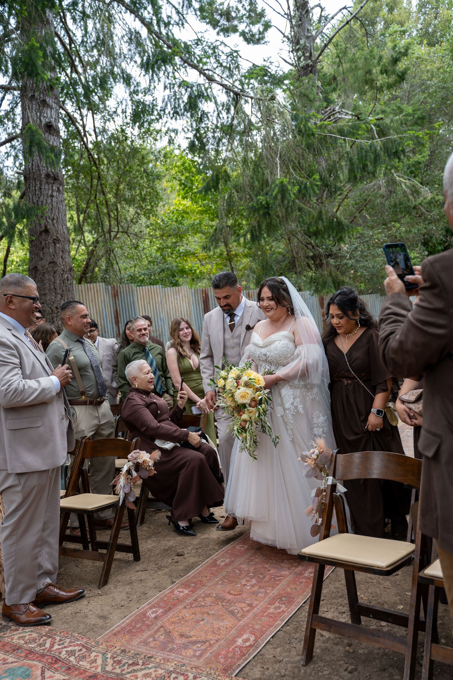 Bride and groom walk down outdoor aisle surrounded by smiling guests at a woodland wedding ceremony.