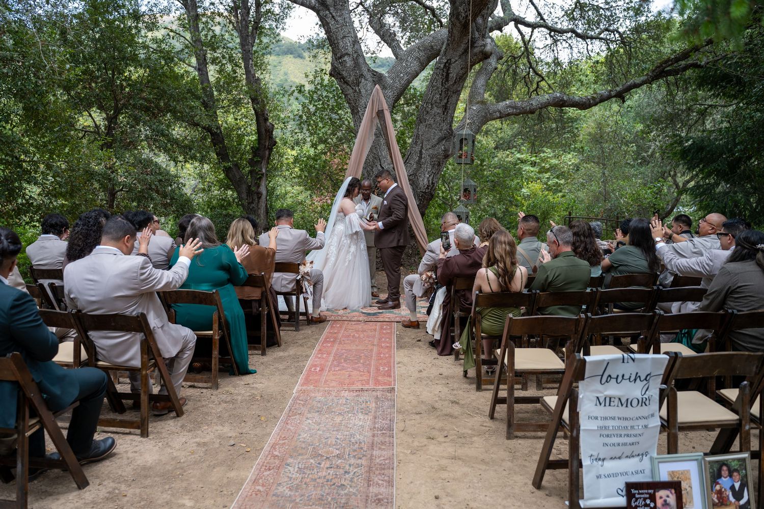 Outdoor wedding ceremony in a wooded setting with guests seated on wooden chairs along a rustic aisle.