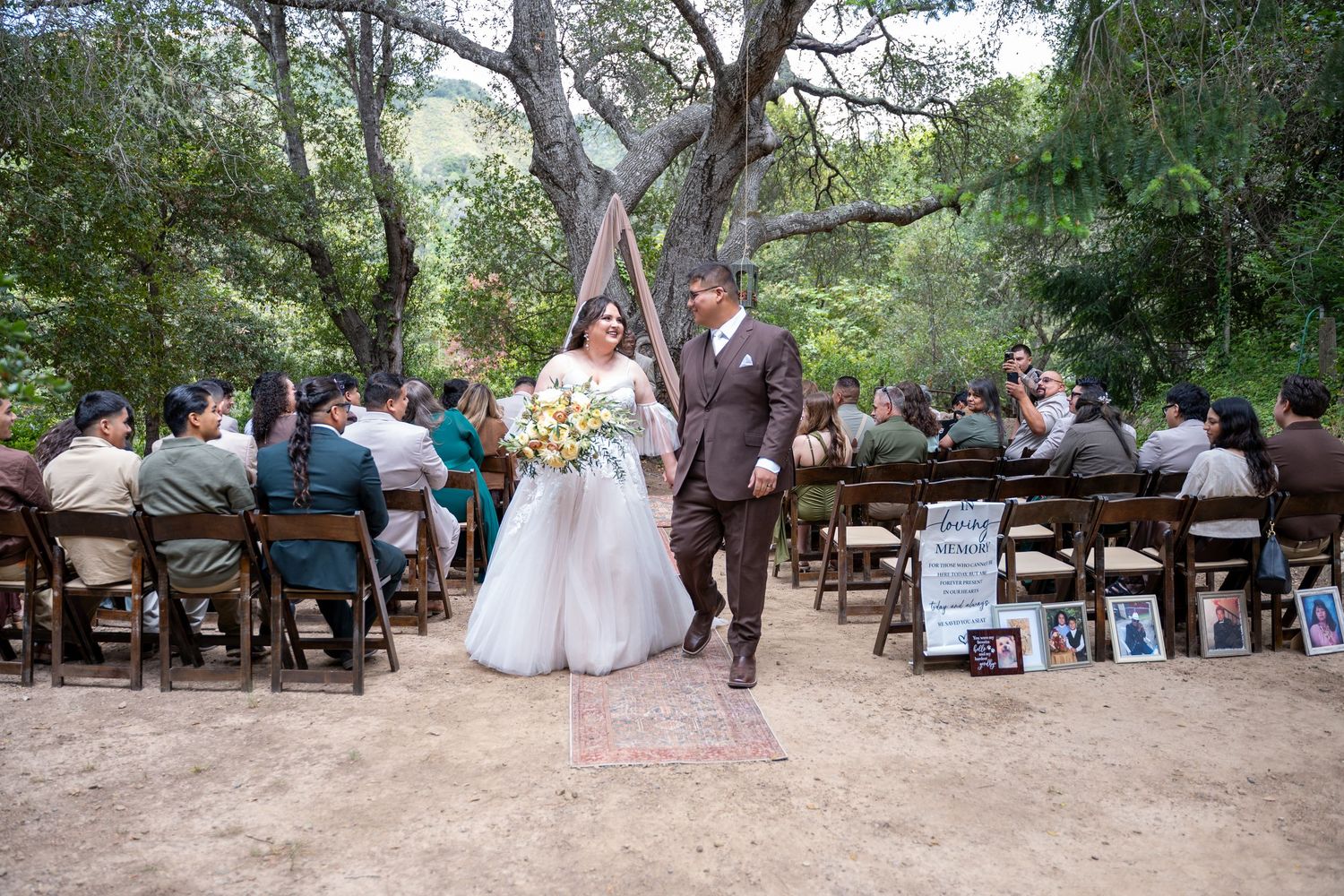 Bride and groom walk down outdoor wedding aisle surrounded by guests seated on wooden chairs under shady trees.