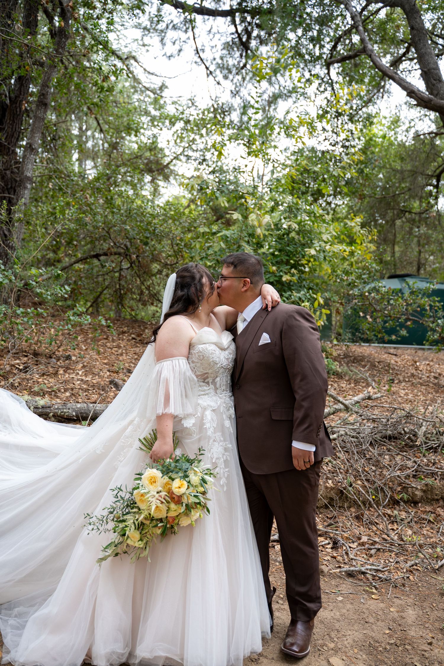 Bride and groom sharing a kiss outdoors in a wooded setting, bride holding yellow floral bouquet in white gown.