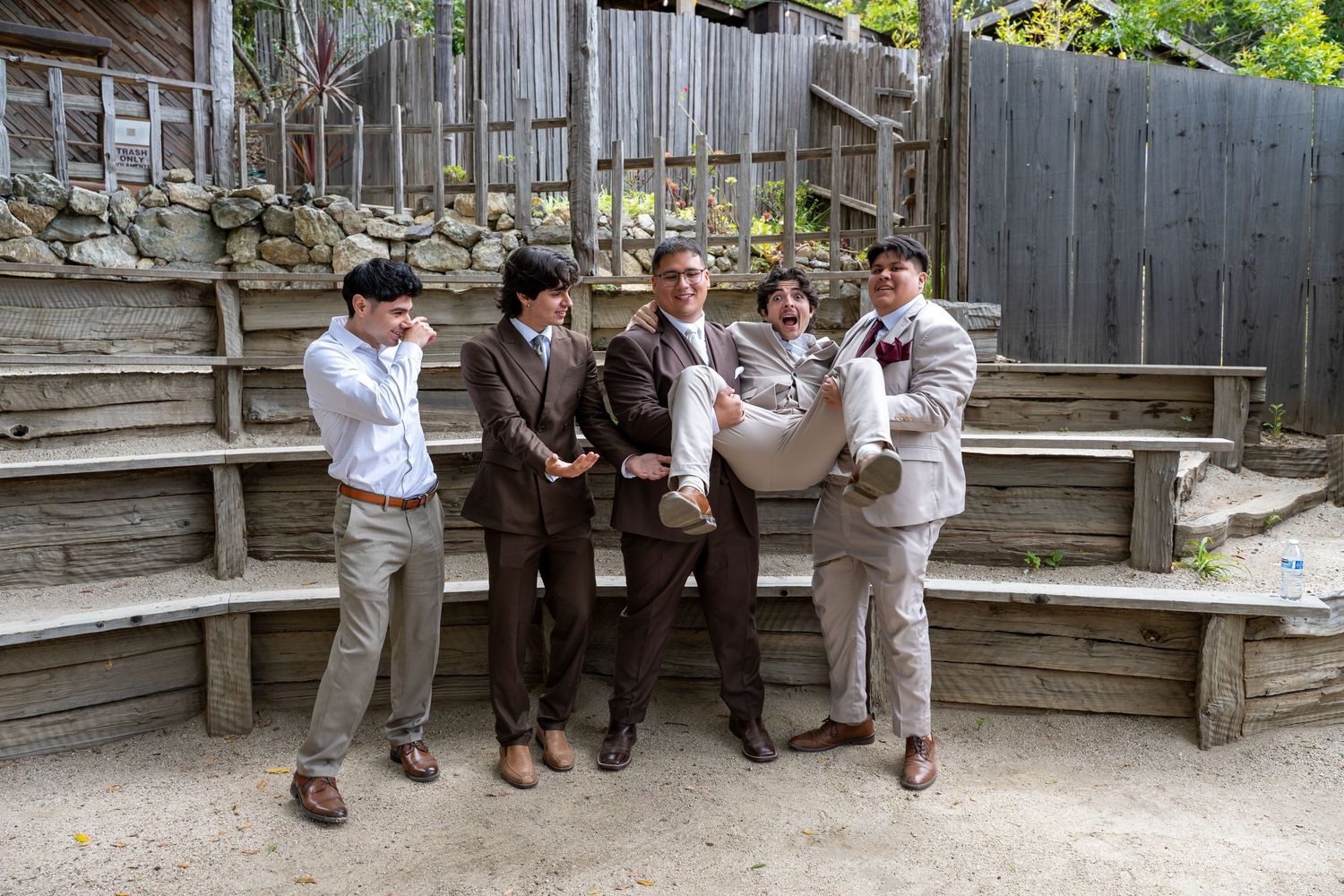 Five groomsmen posing playfully in brown and gray suits at an outdoor rustic venue.