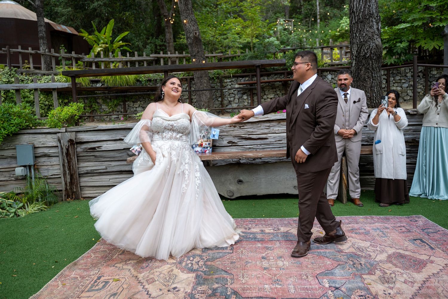 Bride and groom share a joyful first dance at an outdoor woodland wedding reception on a decorative rug.