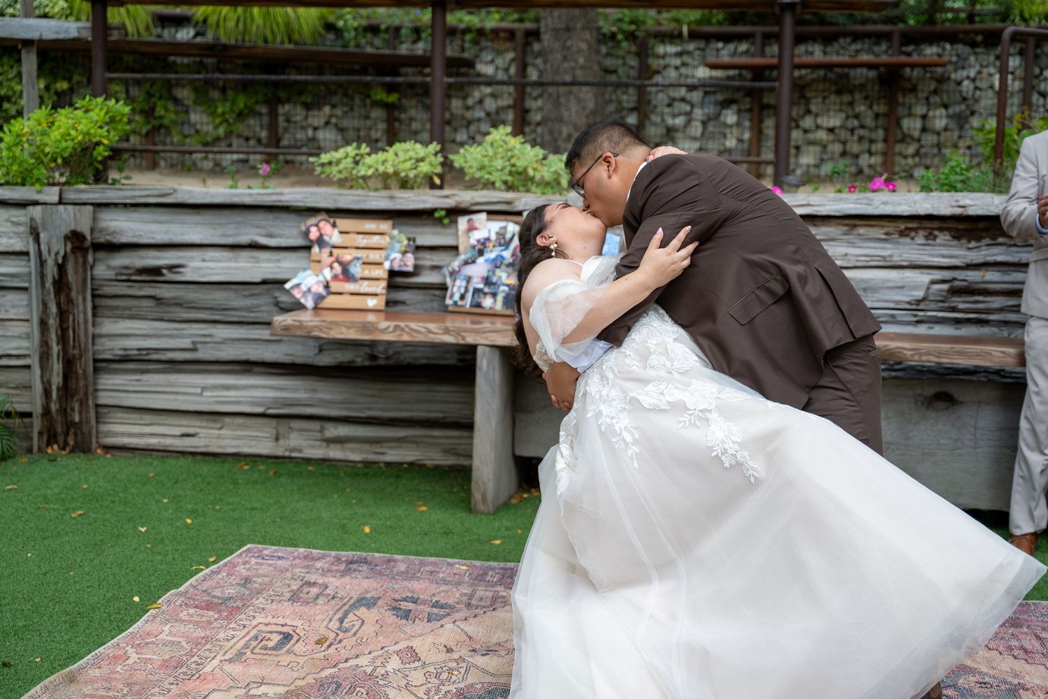 Bride and groom share a romantic kiss at their outdoor rustic wedding venue beside a wooden bench.