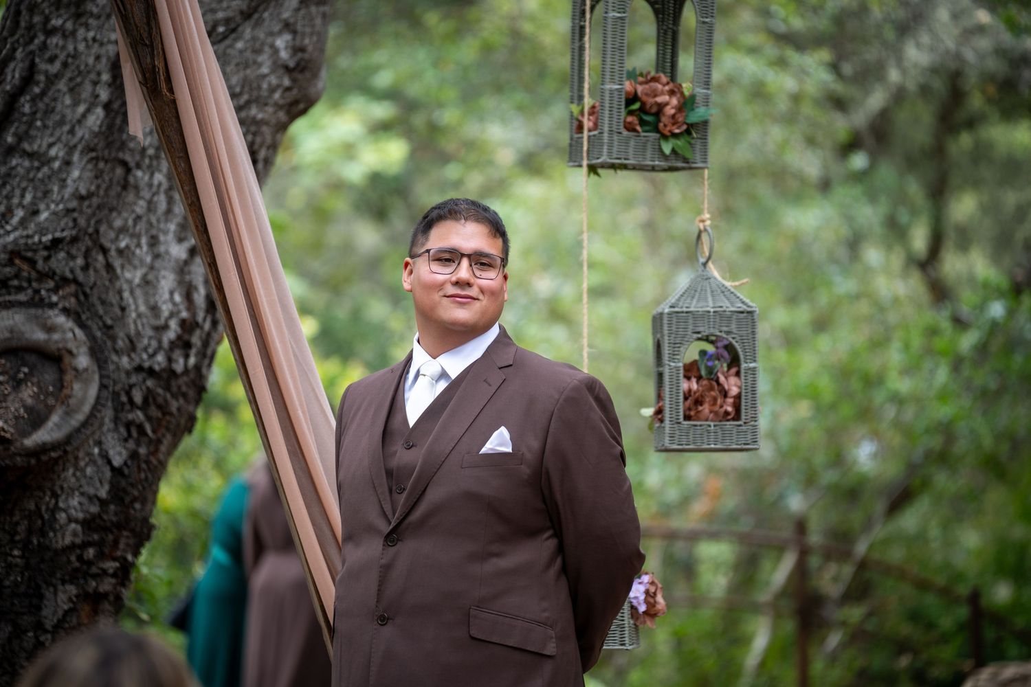 Groom in brown suit stands outdoors at woodland wedding ceremony with hanging lantern decorations among trees.