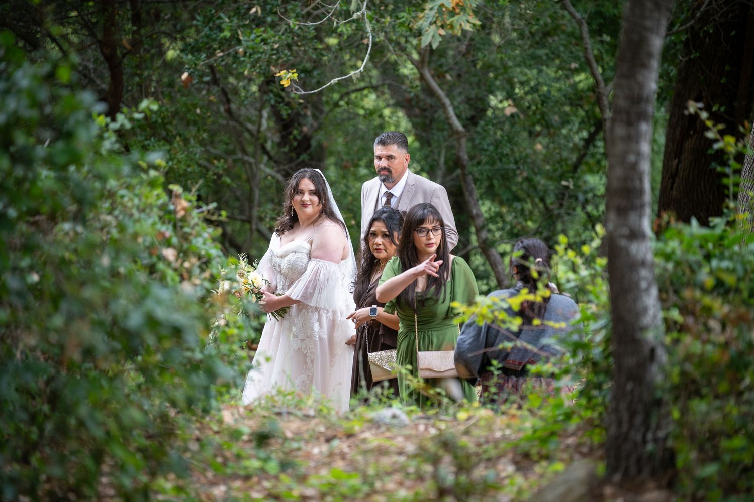 Couple in white dress and suit pose with guests in a lush green forest during an outdoor wedding ceremony.