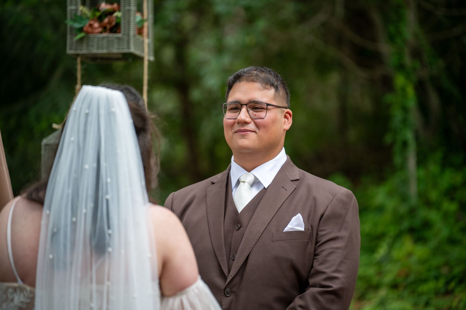 Groom in brown suit smiling at bride during outdoor wedding ceremony in lush green forest setting.