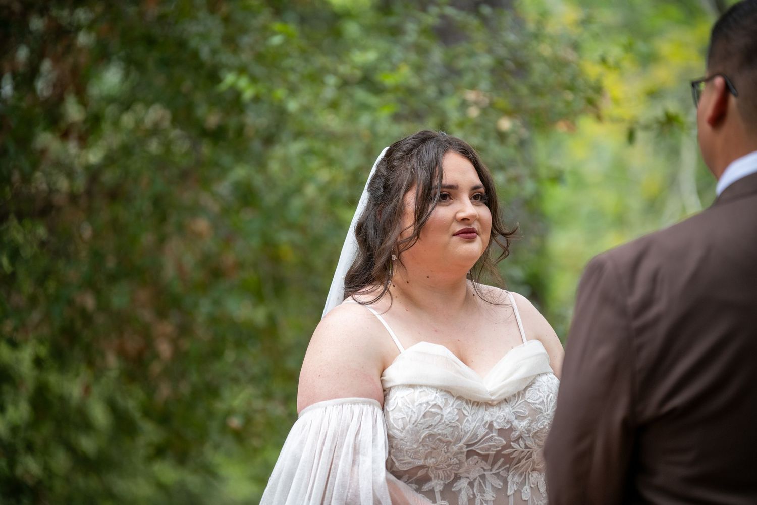 Bride in lace wedding dress and veil gazes emotionally at groom during outdoor wedding ceremony.