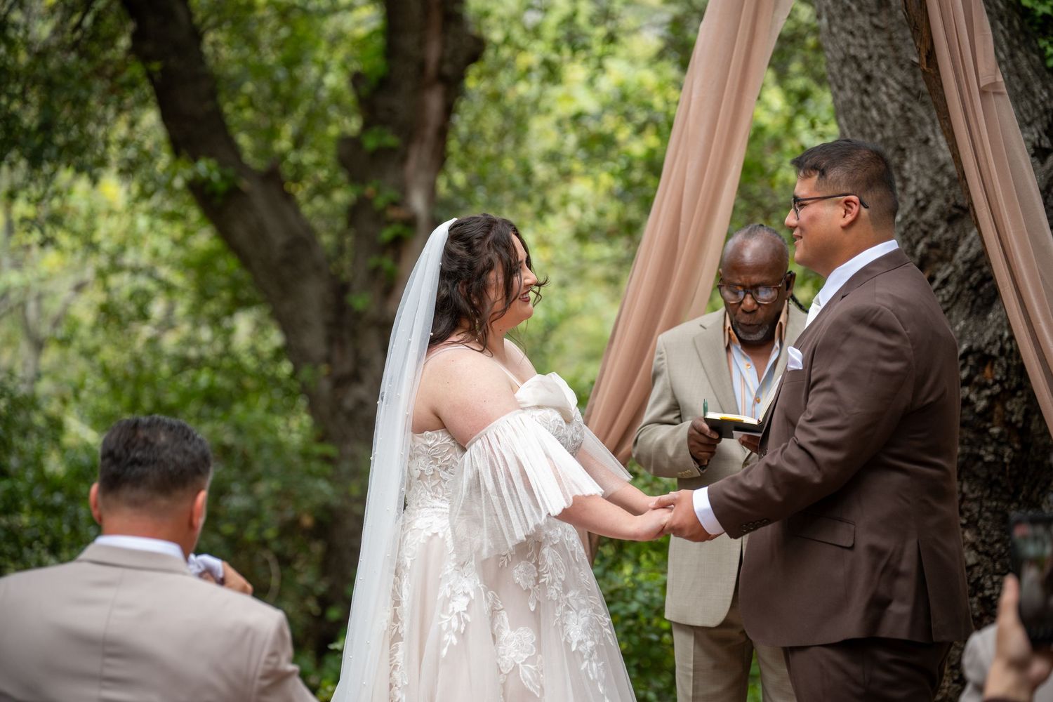 Outdoor wedding ceremony with bride in lace gown and veil holding hands with groom in brown suit before officiant.