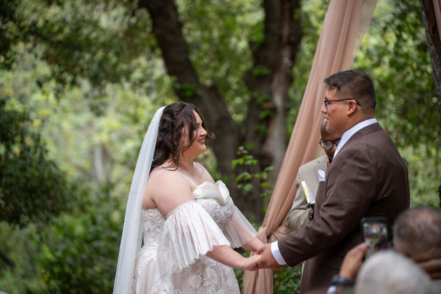 Bride and groom holding hands at outdoor wedding ceremony surrounded by lush green trees with blush pink draping.