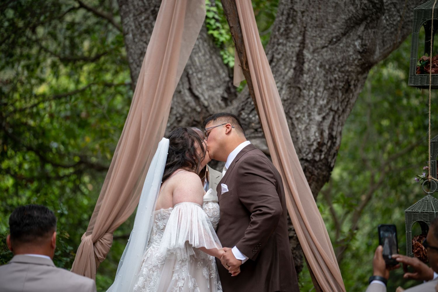 Bride and groom share first kiss at outdoor wedding ceremony under elegant draped arch surrounded by lush greenery.