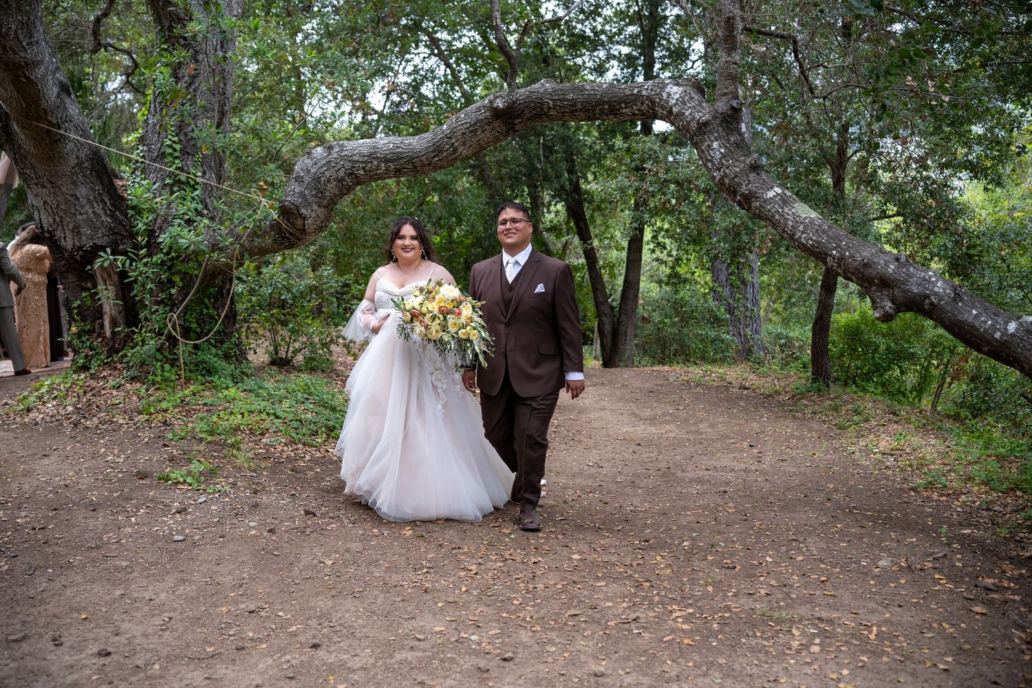 Bride and groom walking hand in hand on a dirt path under arching trees, bride holding a colorful bouquet.