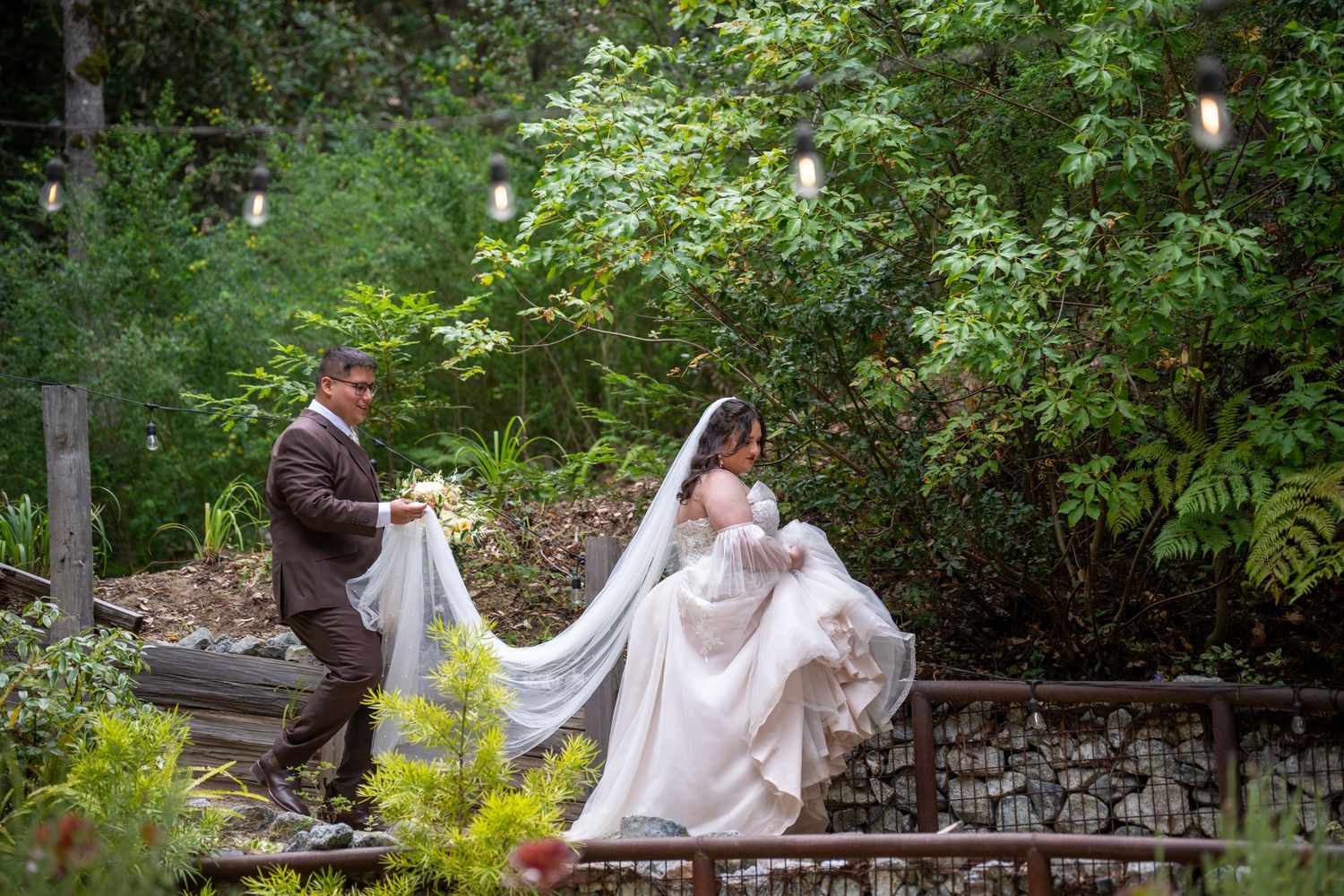 Groom holding bride's veil in lush garden setting during outdoor wedding ceremony with string lights.