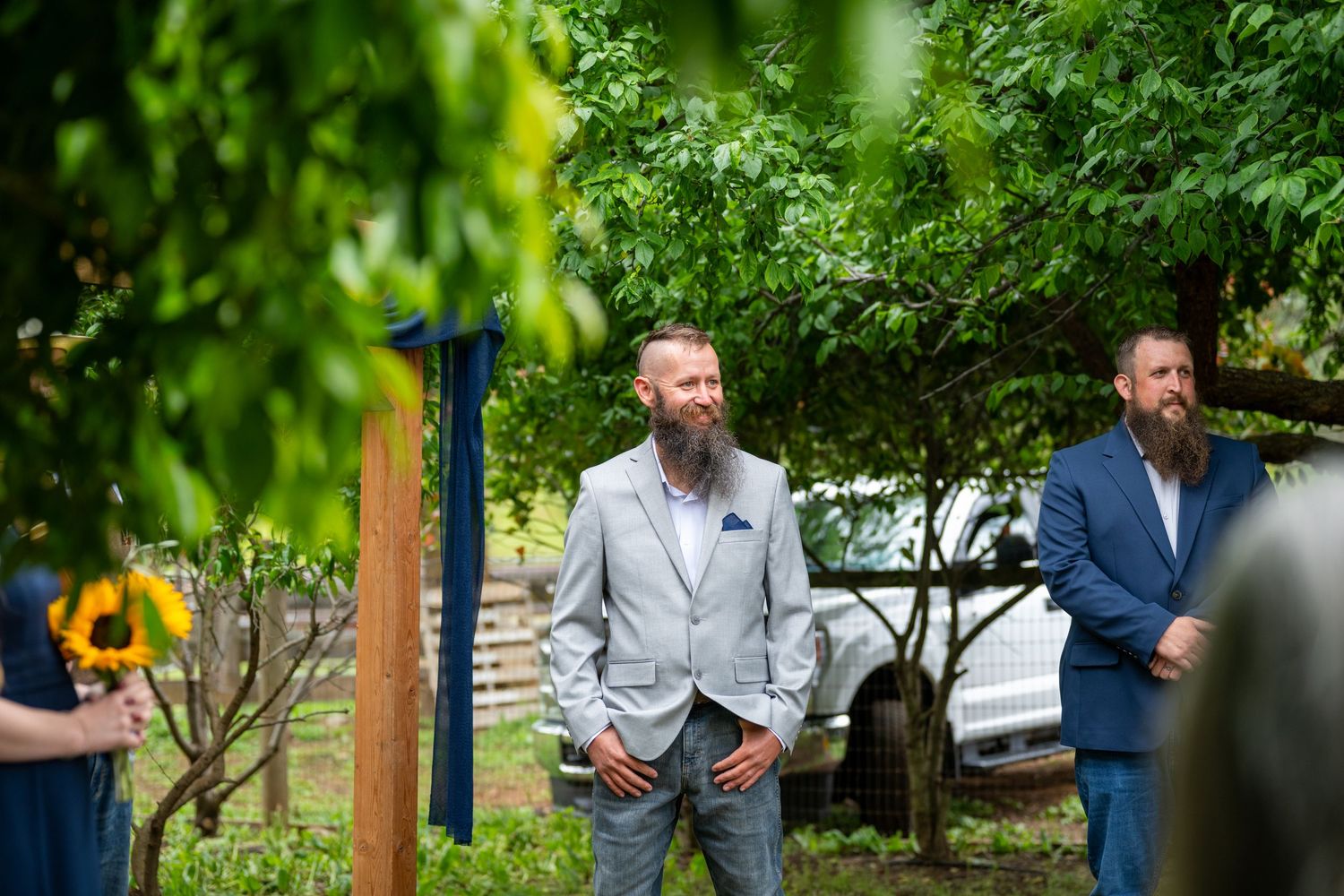 A bearded groom in a gray suit smiles during an outdoor wedding ceremony surrounded by lush green trees.