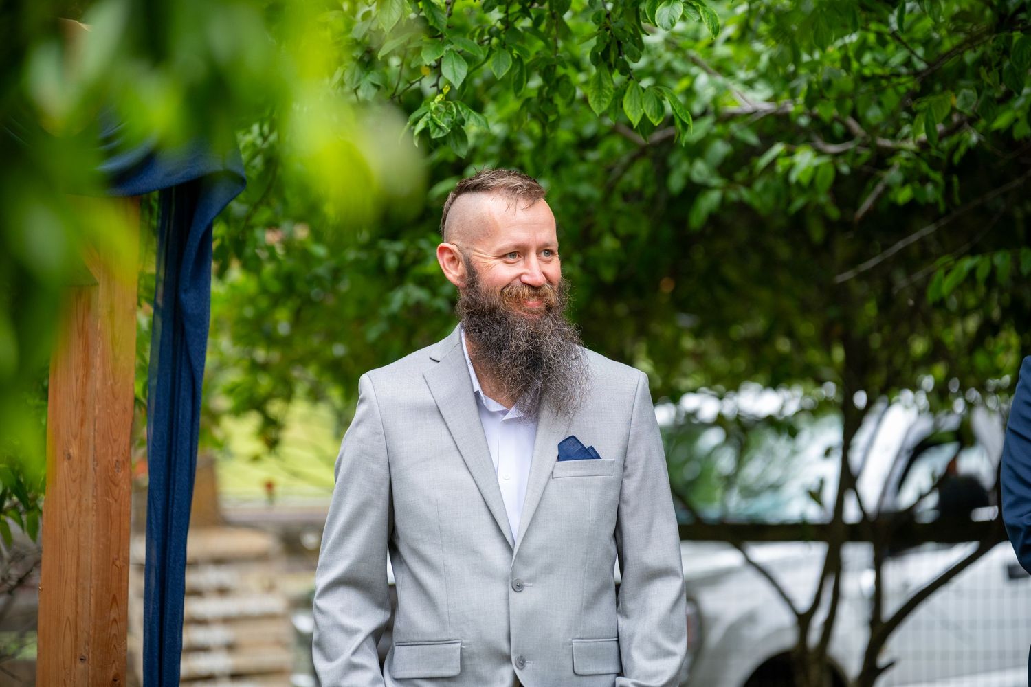 Bearded groom in gray suit smiling outdoors surrounded by lush green foliage before wedding ceremony.