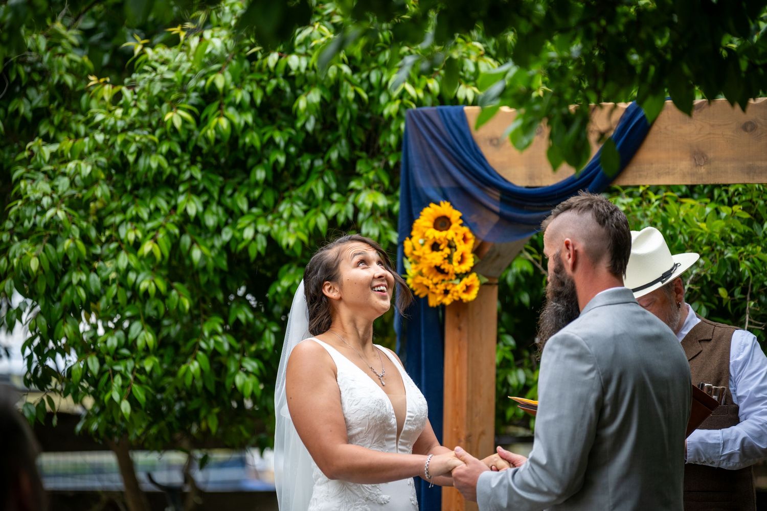 Bride and groom holding hands at outdoor wedding ceremony with sunflower arch and blue draping.