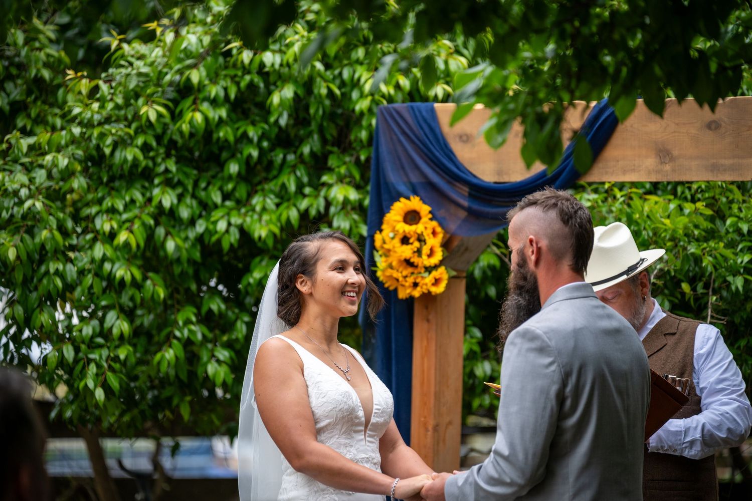 Bride and groom hold hands at outdoor wedding ceremony with sunflower arch and blue drape amid lush green trees.