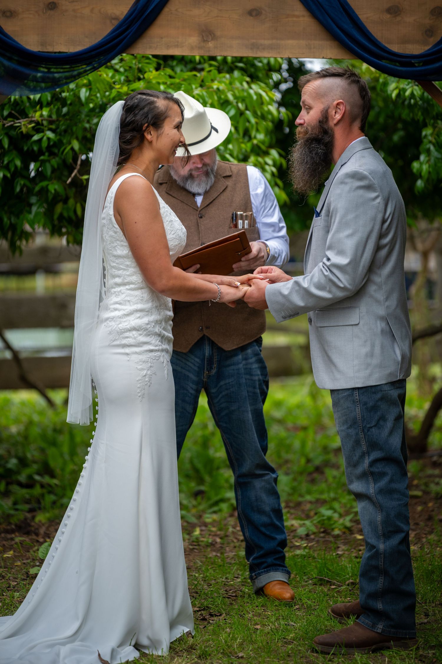 Bride and bearded groom exchange vows outdoors with officiant in cowboy hat at rustic wedding ceremony.