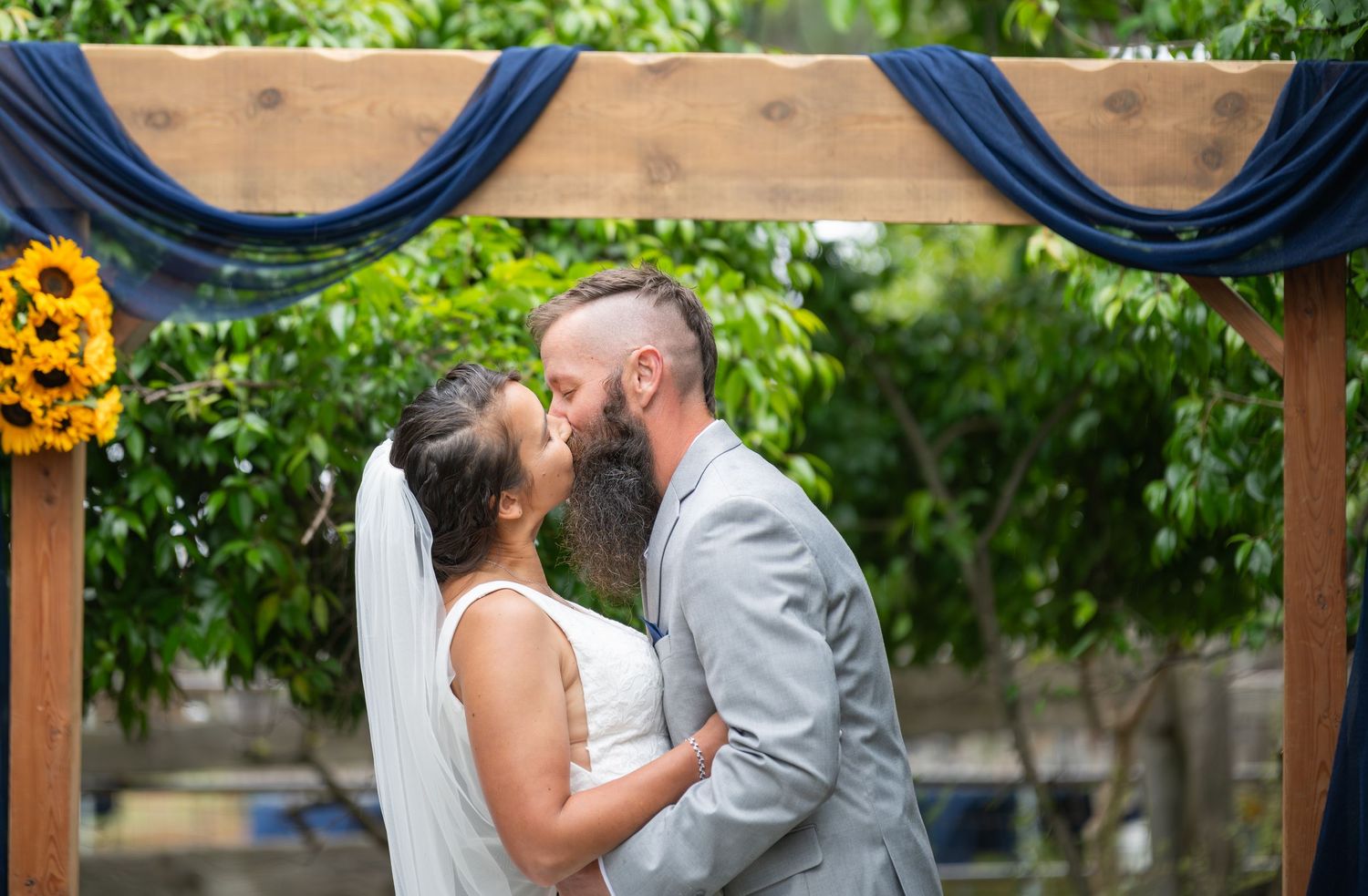 Bride and groom sharing first kiss under rustic wooden arch with navy draping and sunflowers at outdoor wedding ceremony.