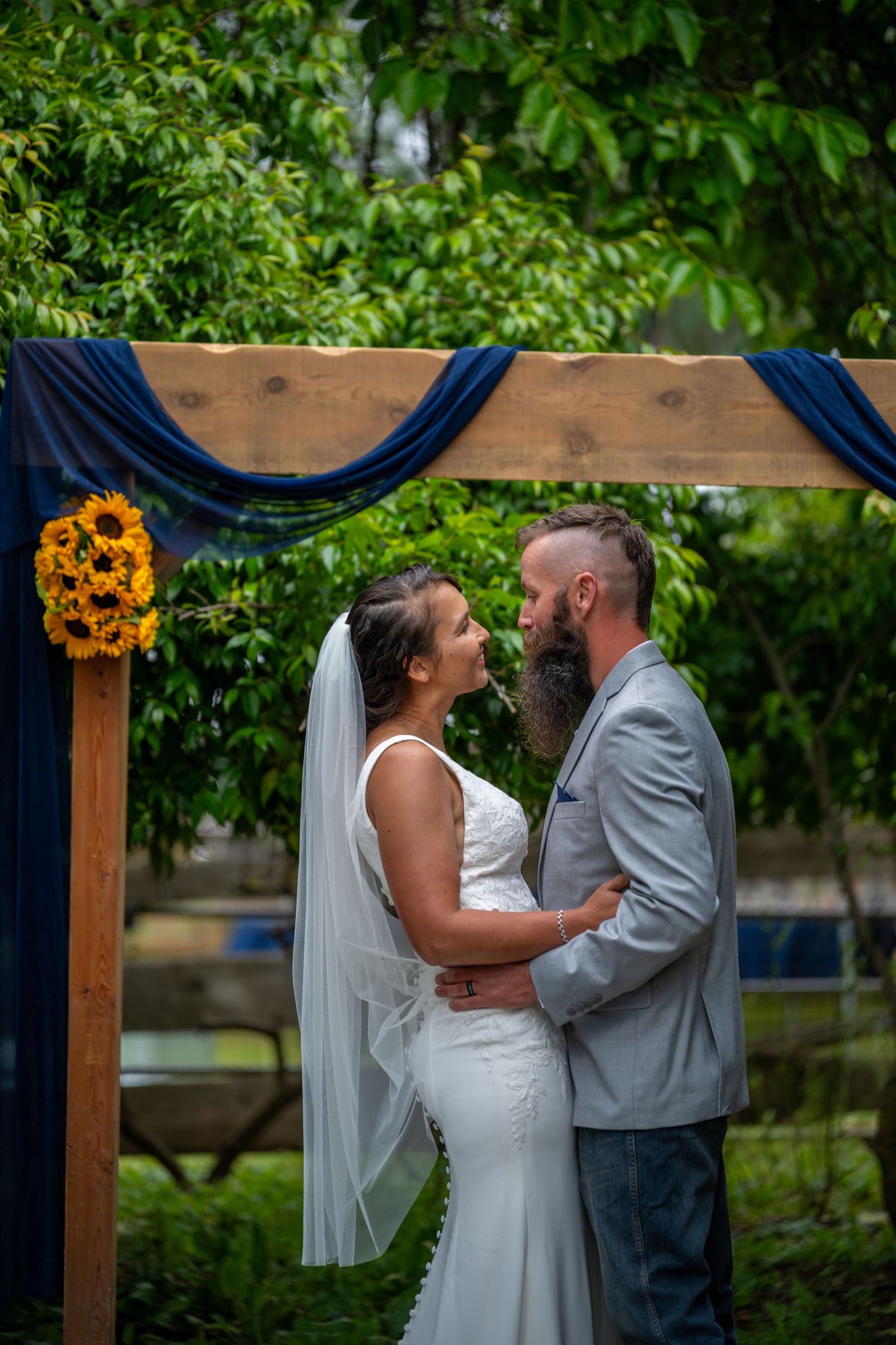 Bride and groom share romantic moment under rustic wooden arch with navy draping and sunflowers at outdoor wedding ceremony.