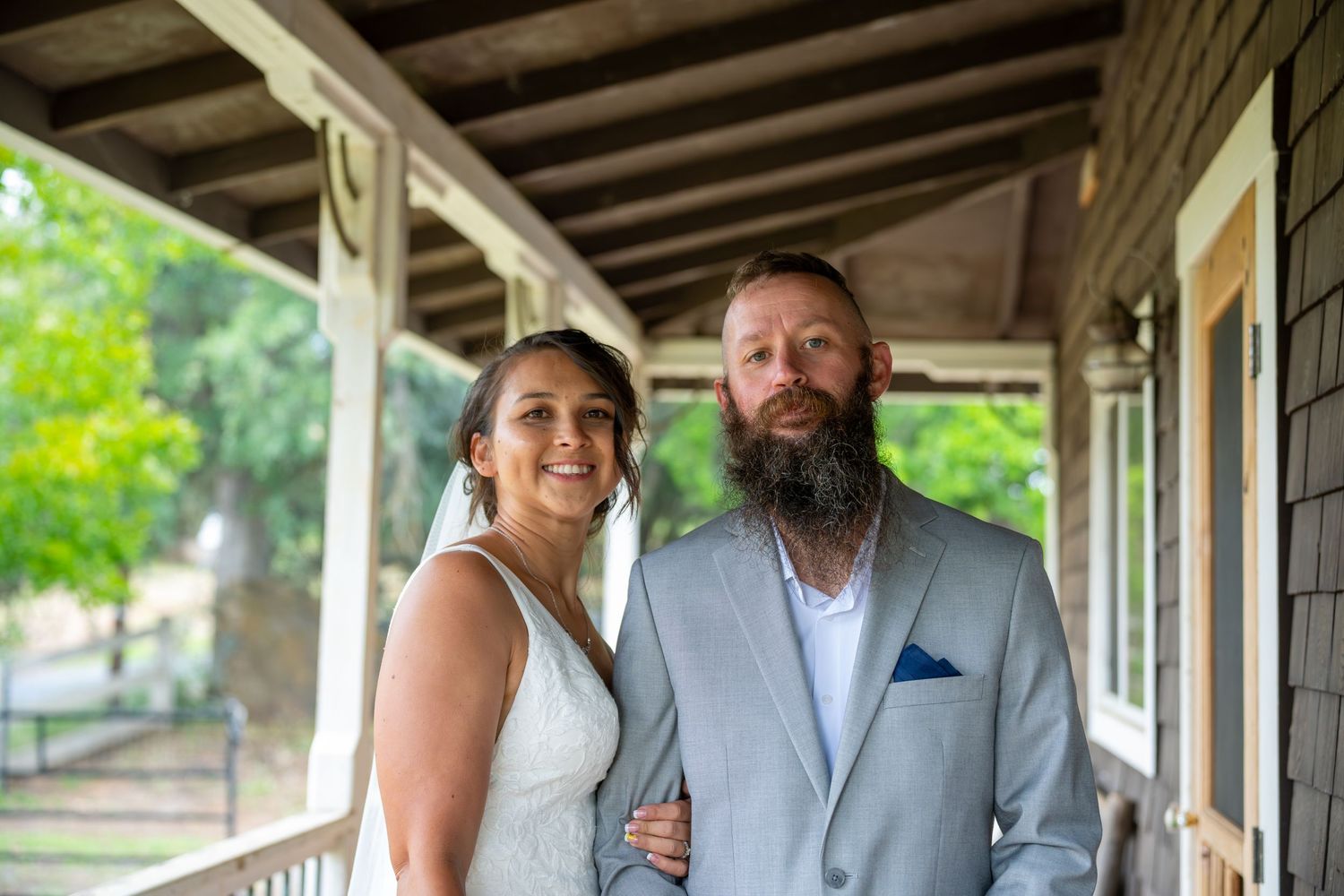 Smiling bride and bearded groom in gray suit pose together on a wooden porch surrounded by greenery.