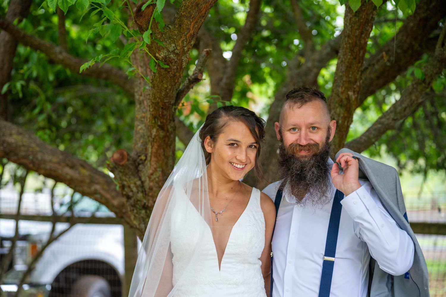 Smiling bride in white gown and veil poses with bearded groom in gray suit under a tree.
