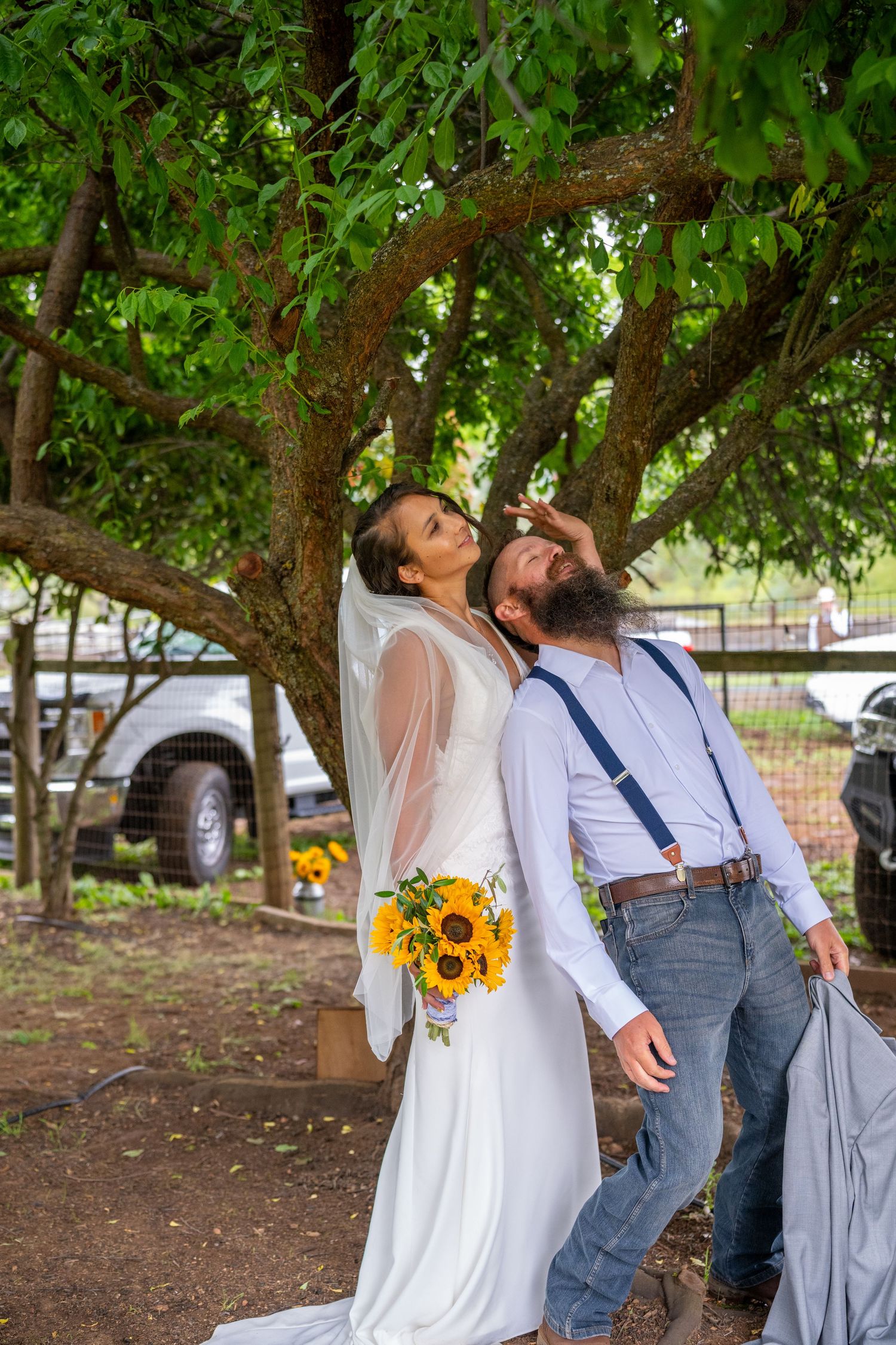 Bride in white gown holding sunflowers and groom in suspenders share a playful moment under a large tree outdoors.