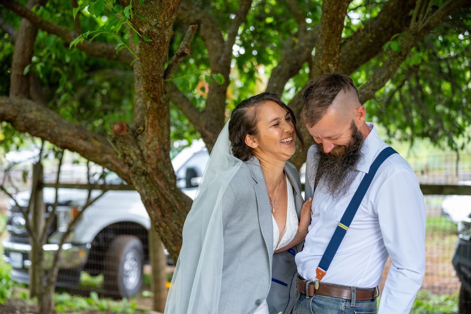 Bride and bearded groom share romantic moment under tree, groom wearing blue suspenders at outdoor wedding.
