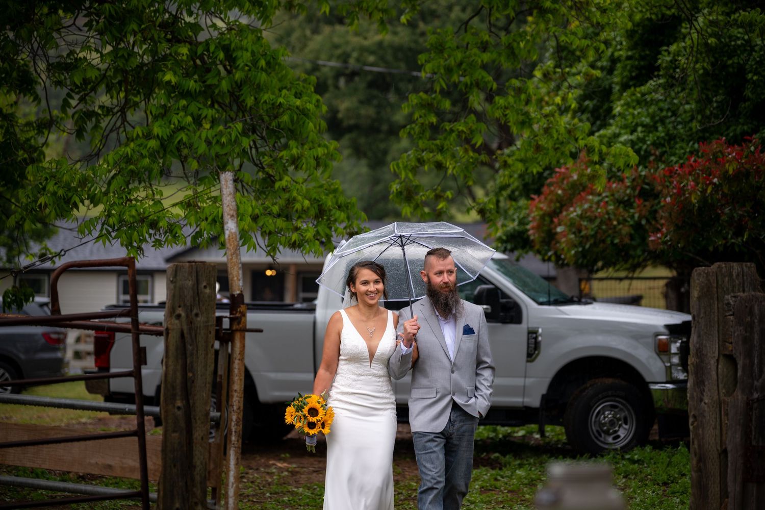Bride and groom walking under umbrella in rain on wedding day with sunflowers and rustic fence backdrop.
