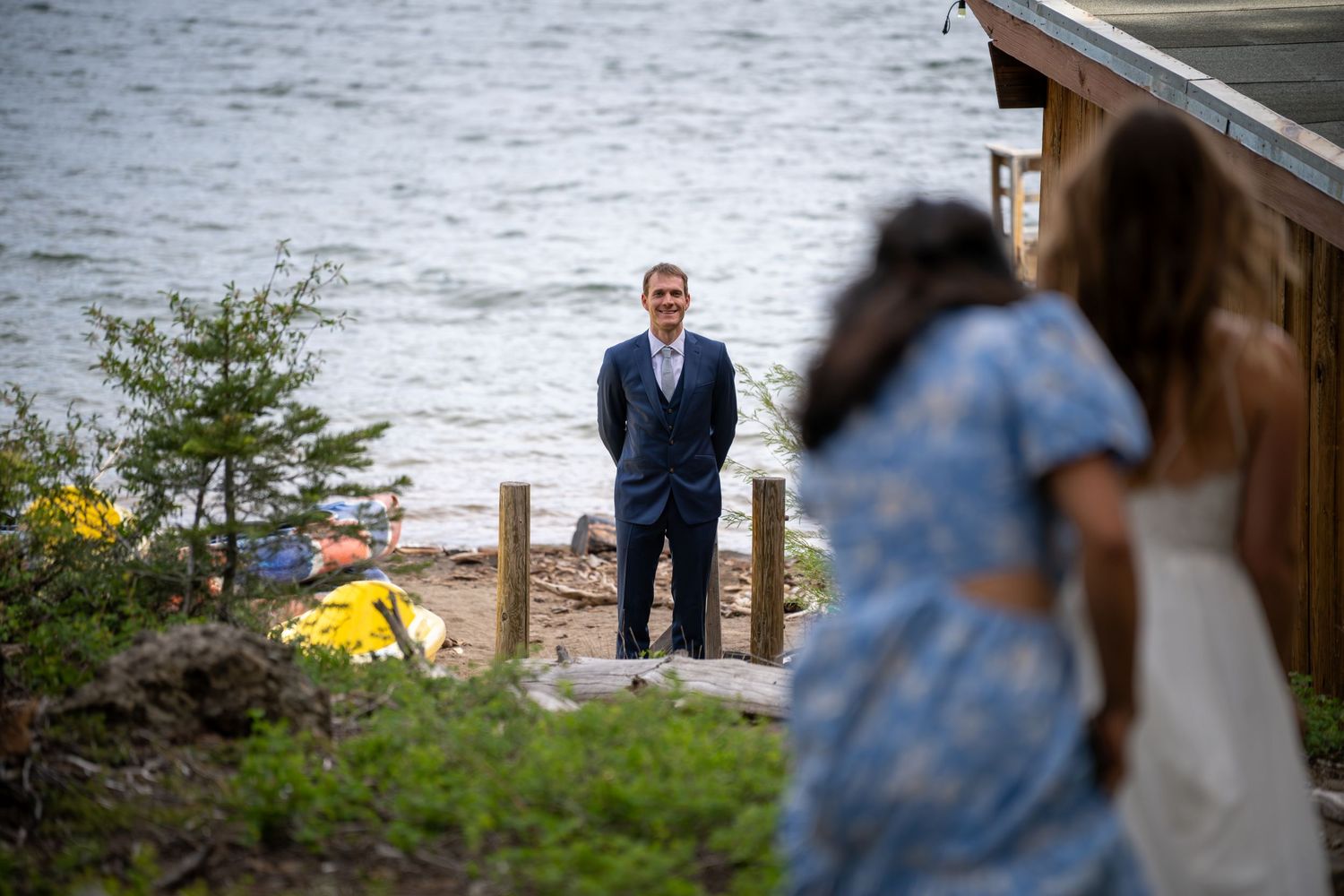 Groom in navy suit stands by lakeside smiling as bride approaches with guest at outdoor waterfront wedding.