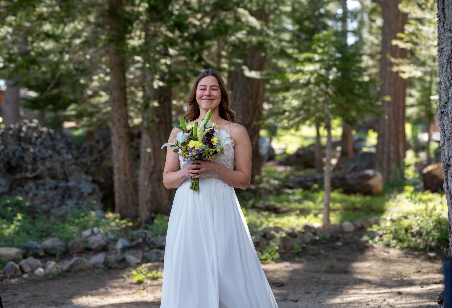 Bride in white gown holding bouquet smiling in forest setting.