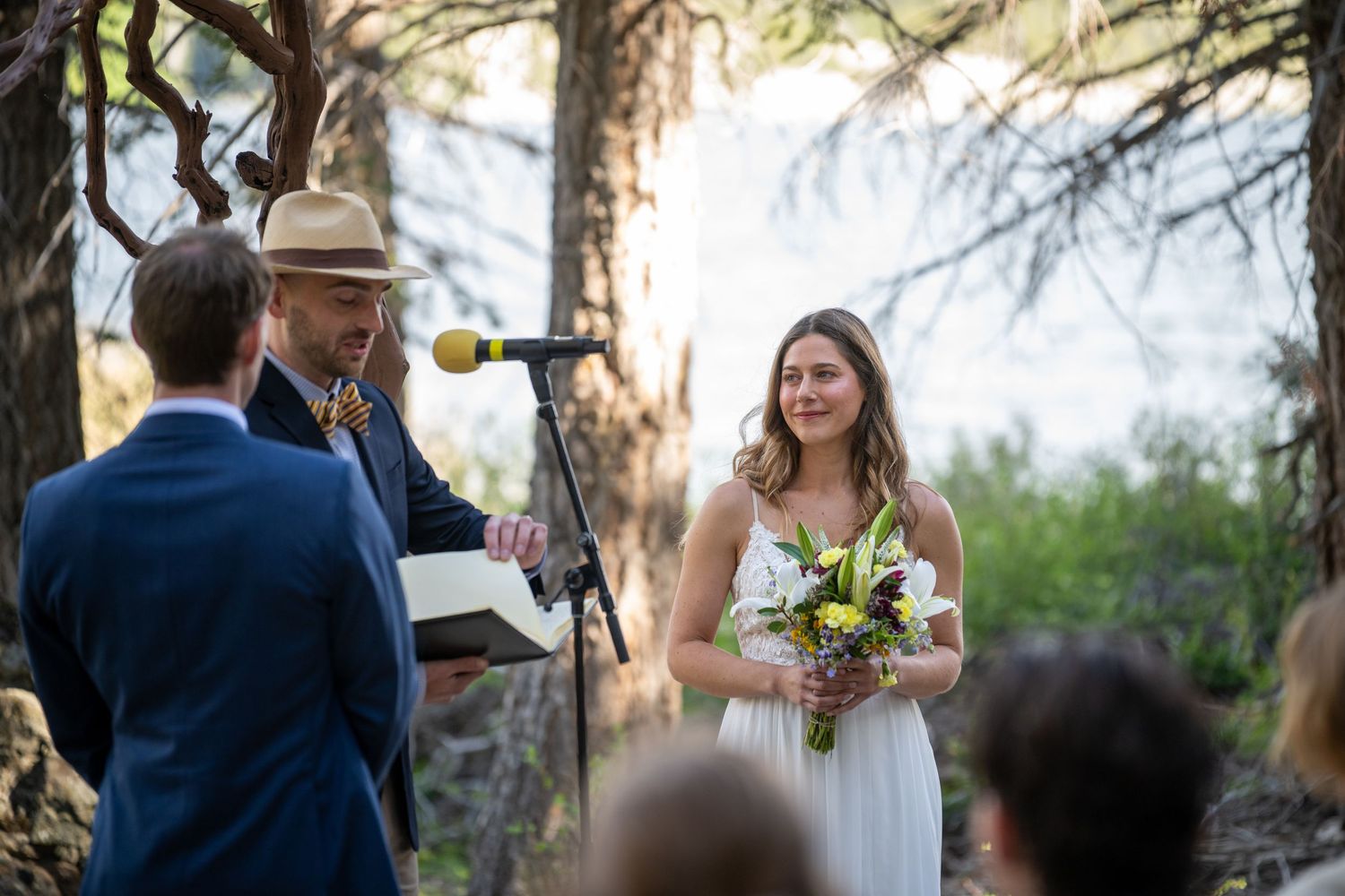 Outdoor wedding ceremony in a forest with a smiling bride holding a floral bouquet facing the groom and officiant.