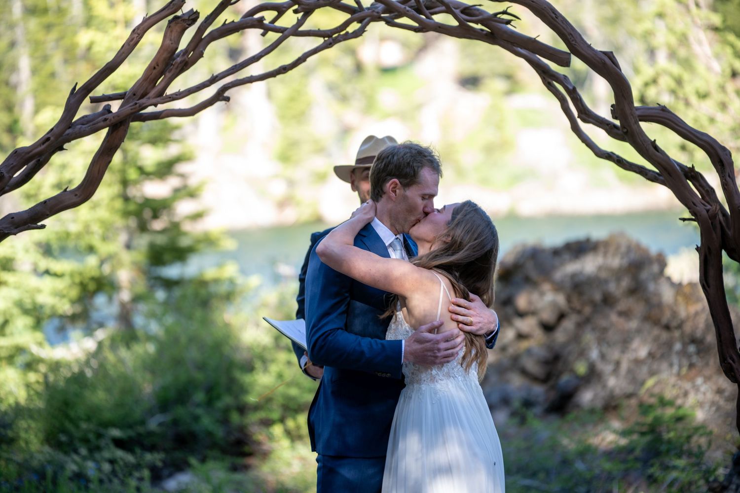 Couple sharing first kiss under rustic tree arch at outdoor mountain wedding ceremony with forest backdrop.