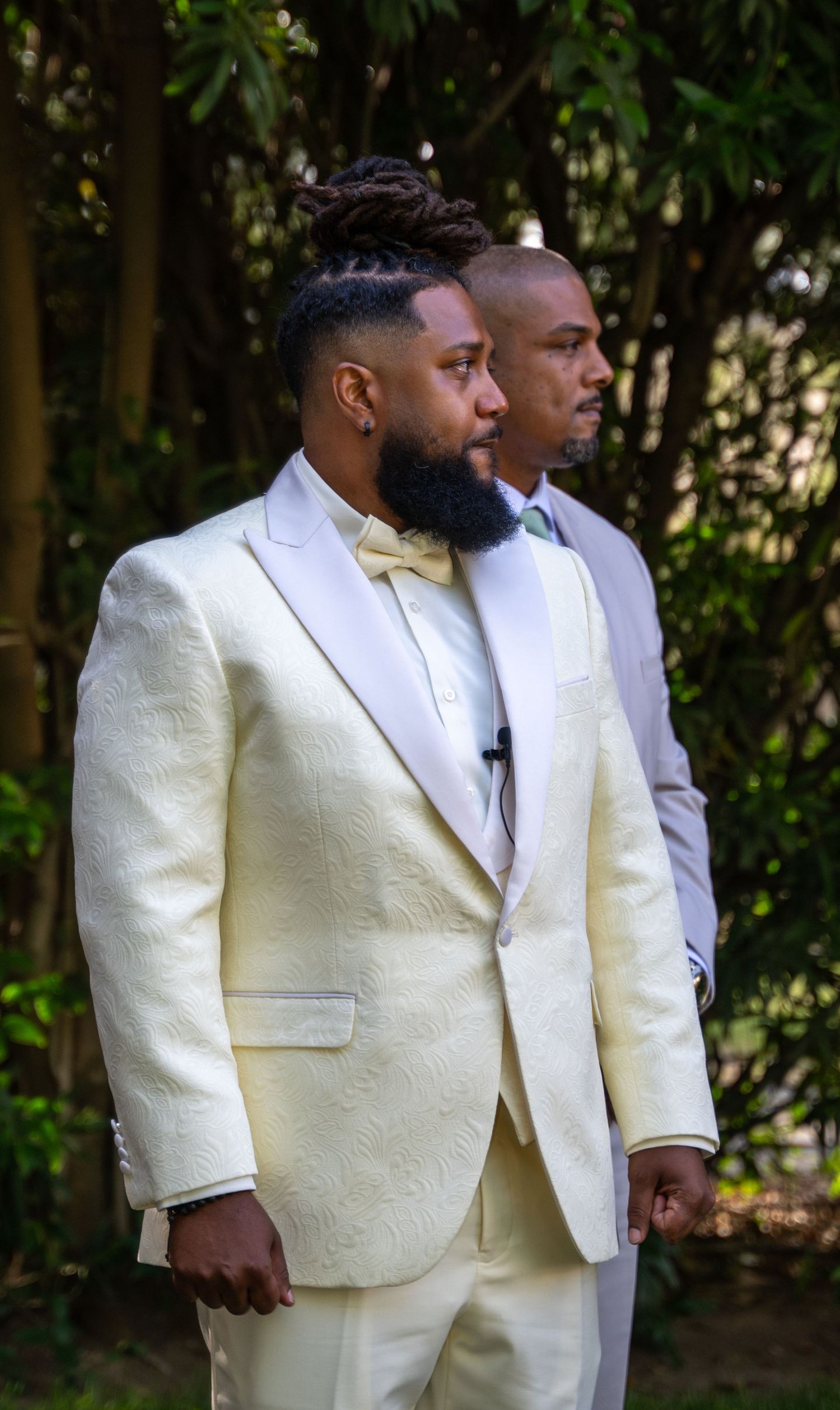 Two men in elegant white tuxedos stand side by side during formal outdoor event.