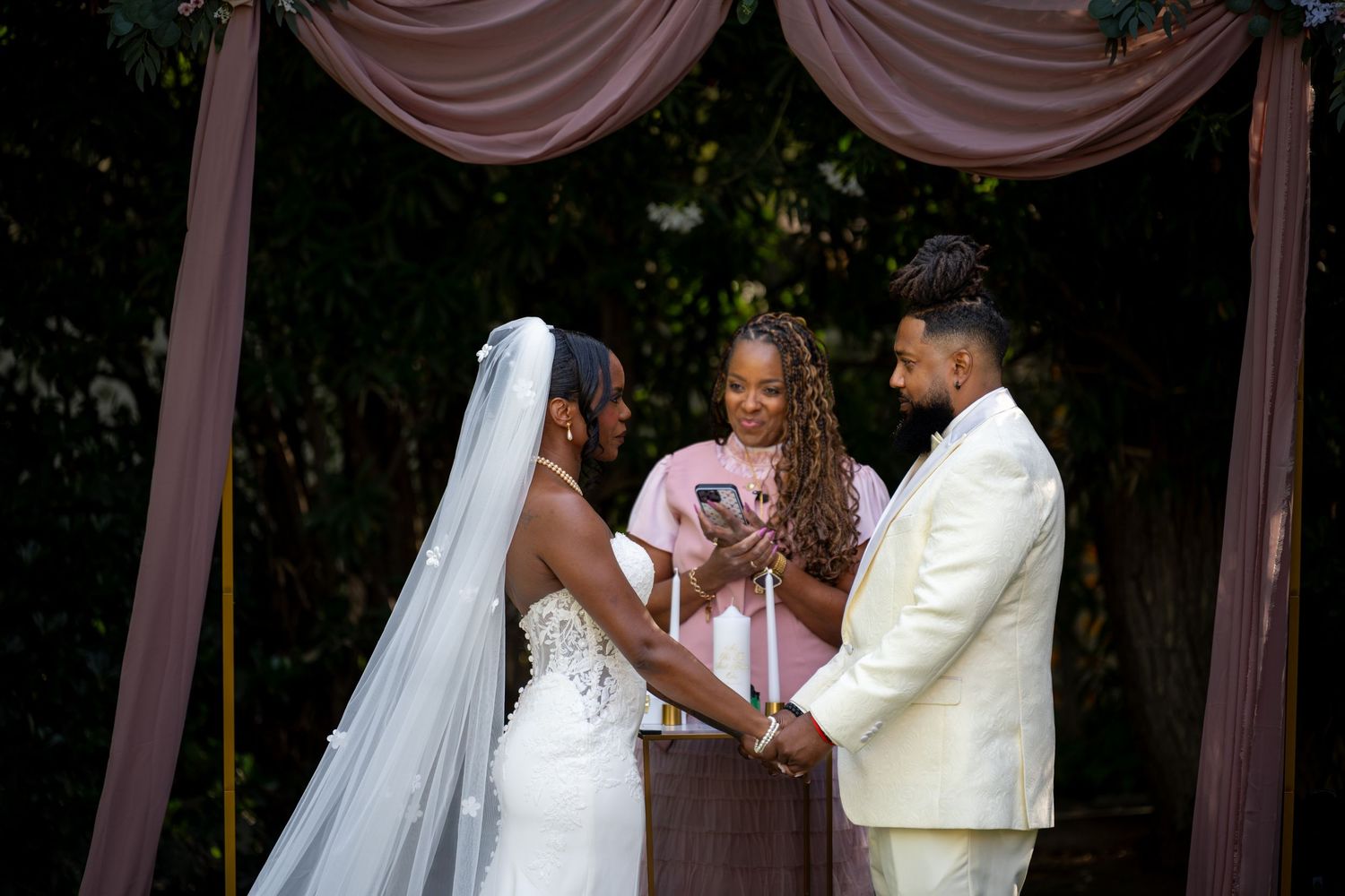 Outdoor wedding ceremony with officiant and couple under pink draped arch.