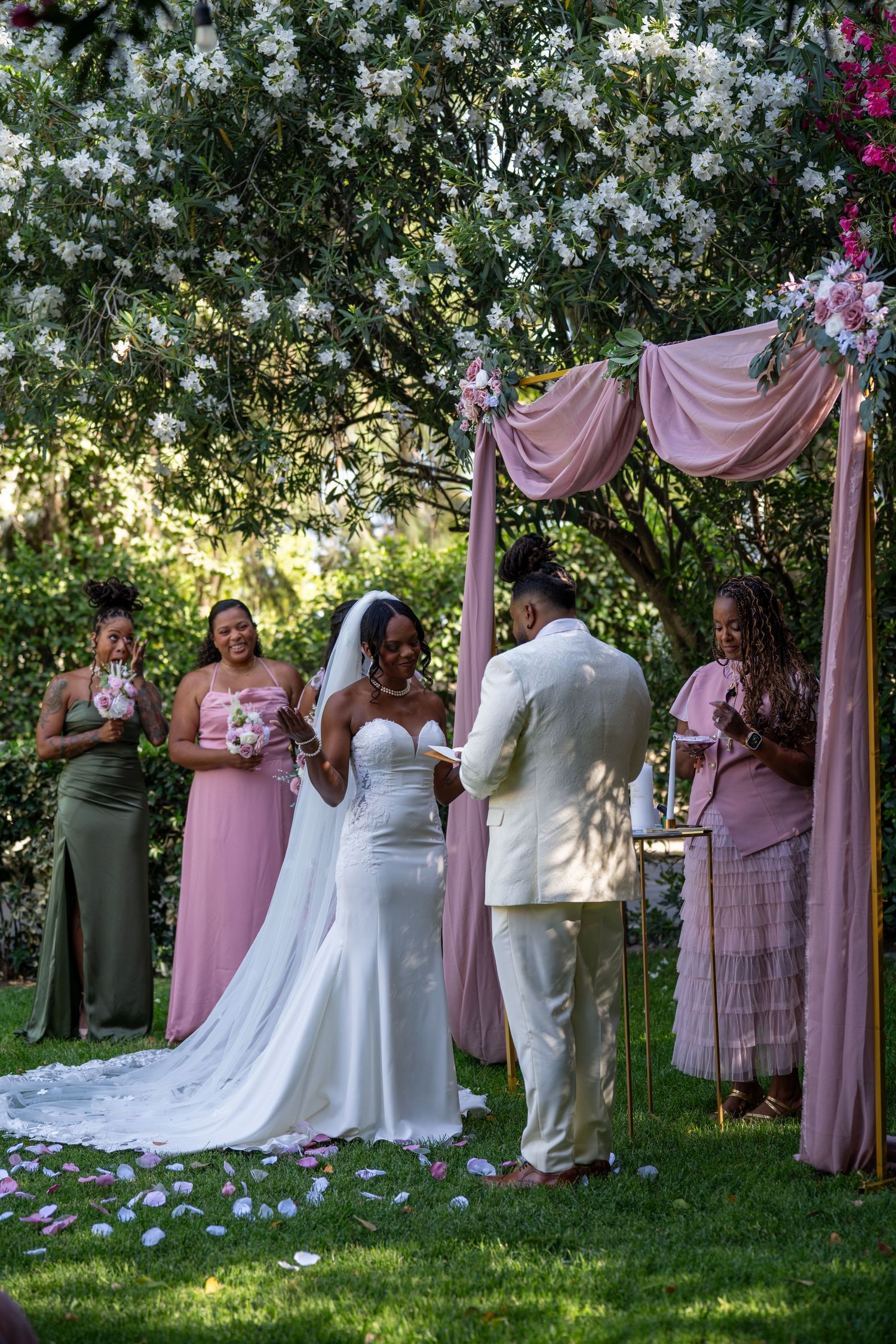 A wedding ceremony in a garden setting with pink drapes and white florals on the grass.